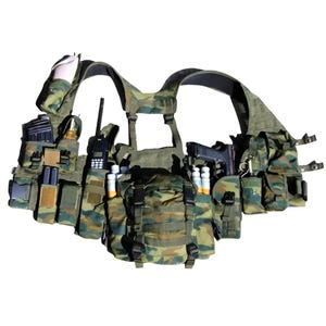 Può includere: Gilet tattico mimetico con più tasche, una radio e una pistola. Il gilet è nei toni del verde, marrone e beige, con vari scomparti per l'attrezzatura. Ha cinghie regolabili.