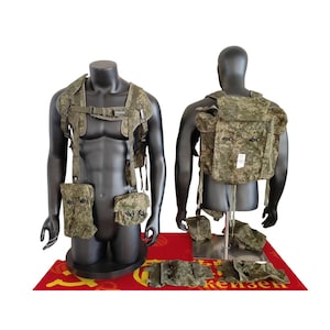 Può includere: Due manichini mostrano giubbotti tattici e tasche in stile militare. I giubbotti e le tasche sono in un motivo mimetico con sfumature di verde e marrone. I manichini sono su una superficie rossa e gialla.