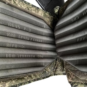 May include: Close-up of a military-style body armor vest. The interior features a gray, ribbed padding with printed text. The exterior has a camouflage pattern. The text includes "6645 P-3" and Cyrillic characters.