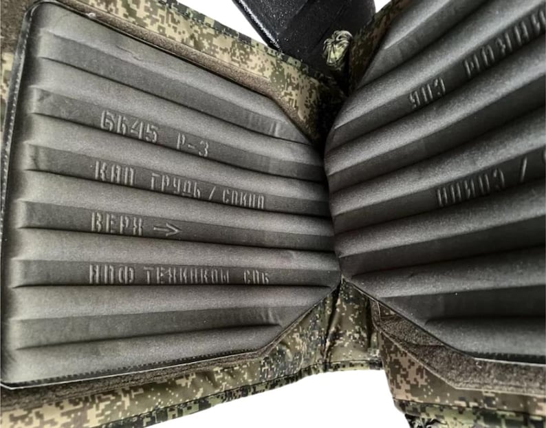 May include: Close-up of a military-style body armor vest. The interior features a gray, ribbed padding with printed text. The exterior has a camouflage pattern. The text includes "6645 P-3" and Cyrillic characters.