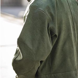 May include: A green jacket with a textured appearance. The jacket features a classic design with a button closure and a structured fit. The sleeves have a curved design at the elbows. The jacket is made of a durable material.