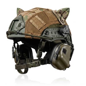 Puede incluir: Un casco táctico verde oliva con una funda de tela de camuflaje y dos orejas de gato puntiagudas en la parte superior. El casco incluye rieles laterales, un soporte frontal, paneles de malla marrón y auriculares de protección verde oliva.