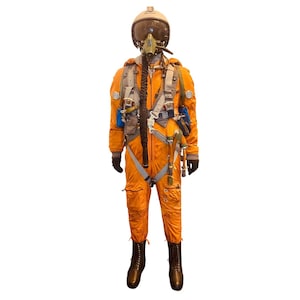 Puede incluir: Un traje de vuelo naranja con casco, máscara de oxígeno y varias correas y equipos. El traje tiene guantes negros y botas marrones. El casco es beige con una visera transparente. La apariencia general sugiere un uniforme de piloto.