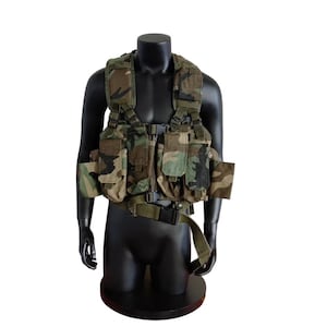 May include: Camouflage tactical vest with multiple pockets and adjustable straps. The vest is displayed on a black mannequin. The vest features a woodland camouflage pattern with shades of green, brown, and black.