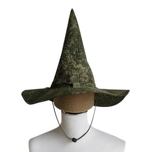 May include: A green camouflage witch hat with a wide brim and a pointed crown. The hat is made of a textured fabric and has a chin strap. The hat is displayed on a mannequin head.