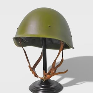 Puede incluir: Casco militar verde oliva con correas de barbilla de cuero, exhibido en un soporte negro. El casco tiene una forma redondeada y está hecho de un material liso y sólido. El soporte tiene una base circular.