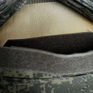 May include: Close-up of a military-style garment, featuring a camouflage pattern in shades of green, gray, and black. The design includes a tan, ribbed interior and a black, textured panel. A dark gray, velcro-like strip is visible.