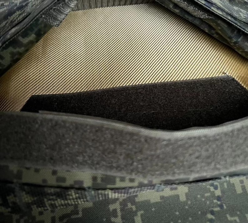 May include: Close-up of a military-style garment, featuring a camouflage pattern in shades of green, gray, and black. The design includes a tan, ribbed interior and a black, textured panel. A dark gray, velcro-like strip is visible.