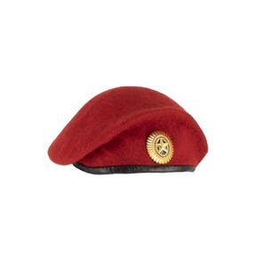 Op de afbeelding: Een donkerrode vilten baret met een zwarte leren rand aan de onderkant. In het midden aan de voorzijde bevindt zich een cirkelvormig, goudkleurig metalen embleem met een sterontwerp en een gestructureerde rand in zonnestraalstijl.