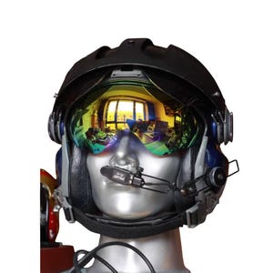 Puede incluir: Casco de piloto negro y azul con visera reflectante. La visera refleja una escena interior colorida. El casco tiene un micrófono y se muestra en una cabeza de maniquí.