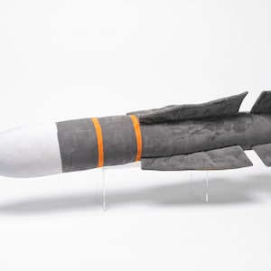 May include: A plush toy shaped like a missile, featuring a white rounded nose cone, a dark grey cylindrical body, and two bright orange bands encircling the midsection. The rear of the object includes four grey stabilising fins.