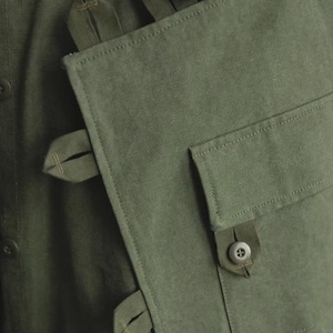 May include: Close-up of a green garment with a unique design. The fabric is a solid olive green, with visible texture. Features include button closures and a flap pocket with a button. The garment has several straps and loops.