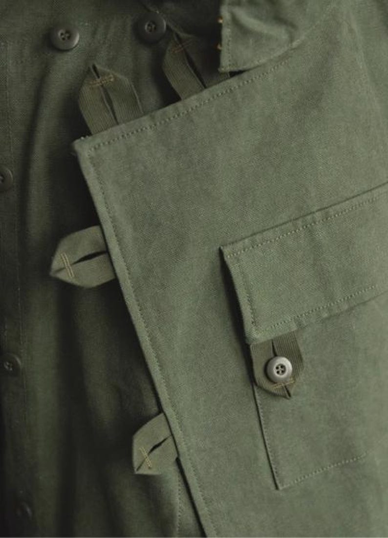 May include: Close-up of a green garment with a unique design. The fabric is a solid olive green, with visible texture. Features include button closures and a flap pocket with a button. The garment has several straps and loops.