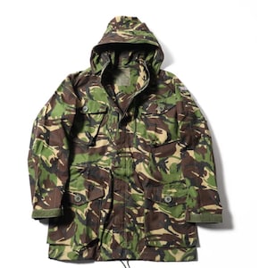 May include: A camouflage jacket with a hood, in shades of green, brown, and beige. The coat has multiple pockets and a drawstring. The military-style jacket is displayed on a white background.