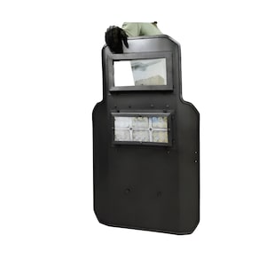 May include: A black ballistic shield with a rectangular viewing window and a smaller rectangular window below. The shield has a curved top and a rounded bottom. A gloved hand is visible at the top.