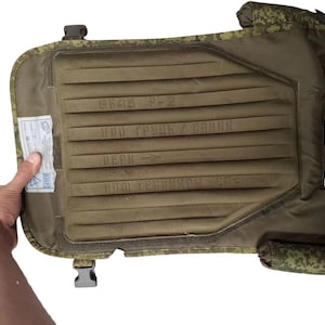 May include: An olive green tactical vest with a padded interior and camouflage trim. The vest has a rectangular design with text and a small tag attached. The vest is designed to be worn on the body.