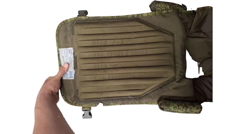 May include: An olive green tactical vest with a padded interior and camouflage trim. The vest has a rectangular design with text and a small tag attached. The vest is designed to be worn on the body.