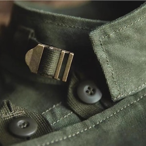 May include: Close-up of an olive green shirt collar with a metal buckle closure. The collar has a button closure and detailed stitching. The fabric appears to be a durable, textured material.