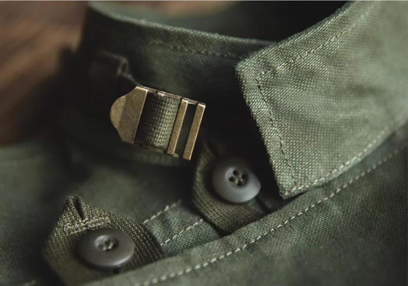 May include: Close-up of an olive green shirt collar with a metal buckle closure. The collar has a button closure and detailed stitching. The fabric appears to be a durable, textured material.