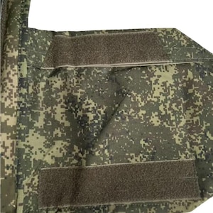 May include: Close-up of a camouflage-patterned fabric with two rectangular hook-and-loop fastener patches. The fabric features a pixelated design in shades of green, brown, and tan. The patches are a muted brown color.