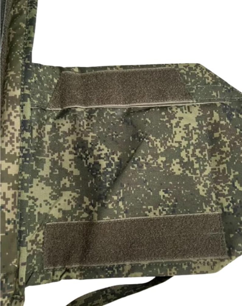 May include: Close-up of a camouflage-patterned fabric with two rectangular hook-and-loop fastener patches. The fabric features a pixelated design in shades of green, brown, and tan. The patches are a muted brown color.