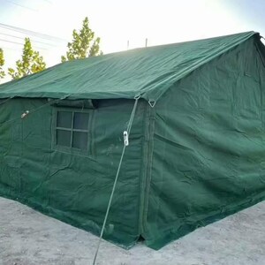 May include: A large, green canvas tent with a pitched roof. The tent has two windows and is secured with ropes and stakes. The tent is set up outdoors on a concrete surface, with trees and a playground visible in the background.