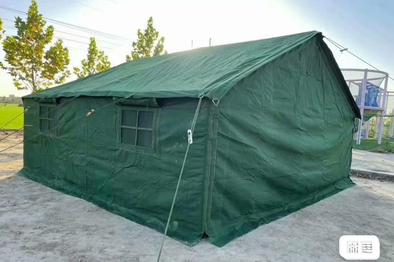 May include: A large, green canvas tent with a pitched roof. The tent has two windows and is secured with ropes and stakes. The tent is set up outdoors on a concrete surface, with trees and a playground visible in the background.