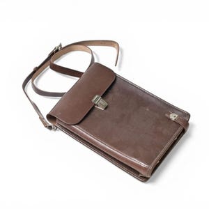 Op de afbeelding: Bruine leren messenger bag met een klepsluiting en schouderriem. De tas heeft een rechthoekige vorm en een metalen sluiting. De verstelbare riem is van bruin leer.