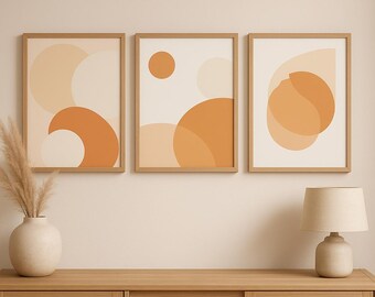 May include: Three framed abstract art prints with geometric shapes in shades of orange and beige. The prints are displayed above a wooden cabinet with a vase of dried plants and a lamp.