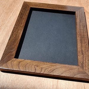 Op de afbeelding: Een rechthoekige houten fotolijst met een donkerbruine afwerking. De lijst heeft een zwart interieur en staat op een lichtbruin houten oppervlak. De houtnerf van de lijst is zichtbaar.