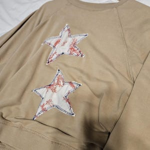 May include: A tan long-sleeve sweatshirt featuring two star-shaped fabric appliqués on the front. The stars are white with a red floral pattern and are attached with visible dark blue stitching. The sweatshirt has a crew neckline and ribbed cuffs.