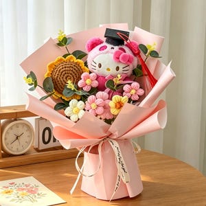 Puede incluir: Un ramo rosa con un Hello Kitty de peluche con birrete, rodeado de flores de ganchillo y un girasol. El ramo está envuelto en papel rosa y atado con una cinta.