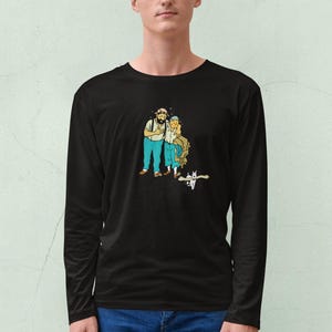 May include: Black long-sleeve shirt featuring a cartoon illustration of two figures with beards and hats, standing together. The figures are wearing blue pants and white shirts. A small cat is at their feet. The shirt is made of a soft material.