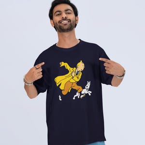 May include: Navy blue t-shirt featuring a cartoon illustration of a character in a yellow coat and brown pants, running with a white dog. The graphic is centered on the shirt.