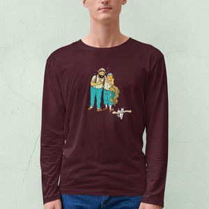 May include: A maroon long-sleeve shirt featuring a cartoon illustration of two people and a cat. The cartoon characters are wearing blue pants and white shirts. The cat is white with black markings. The shirt is made of a soft material.