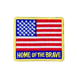 Könnte beinhalten: Gestickter Aufnäher mit dem Design der amerikanischen Flagge, mit roten und weißen Streifen, blauen Sternen und dem Text "HOME OF THE BRAVE" in Weiß auf blauem Grund. Der Aufnäher hat einen gelben Rand.