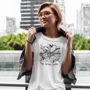 Puede incluir: Camiseta blanca con un diseño de mapa abstracto en blanco y negro y el texto "The Urban Web". La camiseta se lleva con una chaqueta vaquera y gafas.