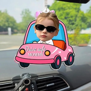 Könnte beinhalten: Ein rosa, auto-förmiger Autoanhänger mit dem Gesicht eines Kindes mit Sonnenbrille. Das Auto hat schwarze Räder, eine blaue Windschutzscheibe und den Text "Drive safe MOMMY". Der Anhänger hängt an einer silbernen Kette.