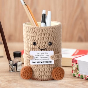 May include: A tan crochet pencil holder shaped like a potato with a smiling face and small arms and feet. The holder has a small white card that reads "I may be a tiny potato, but I can tell - You are an amazing person! YOU ARE AWESOME!" Several pens and pencils are inside.