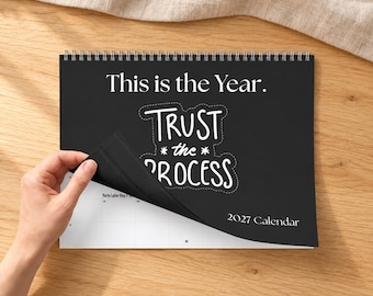 May include: A black 2027 wall calendar with white text that reads This is the Year. Trust the Process. The calendar is spiral-bound at the top and features a white grid page partially visible underneath the black cover page.