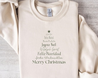 May include: A sand-colored crewneck sweatshirt featuring a dark green graphic of a Christmas tree formed by the words God Jul, Feliz Natal, Buon Natale, Joyeux Noël, Wesołych Świąt, Feliz Navidad, Frohe Weihnachten, and Merry Christmas.