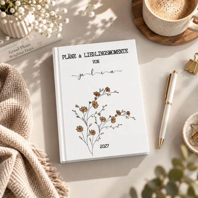 May include: A white hardcover planner with the text PL&Auml;NE & LIEBLINGSMOMENTE VON julia and the year 2027 printed on the front cover. The cover features a black line art illustration of a floral branch with small brown-colored flower petals.