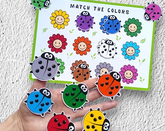 以下が含まれることがあります： 「MATCH THE COLORS」の文字と、カラフルなテントウムシと花のイラストが描かれた白いボードを使った、色のマッチング教育ゲーム。赤、黄色、緑など、さまざまな色のテントウムシの切り抜きが含まれています。