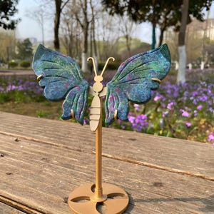 May include: A decorative butterfly sculpture with blue and green wings, accented with pink and gold. The butterfly is mounted on a gold-colored stand with a circular base. The sculpture is displayed outdoors on a wooden surface.
