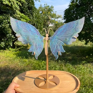 Peut inclure: Un présentoir à bijoux en forme de papillon avec deux grandes ailes texturées dans des tons de bleu, violet et sarcelle. Les ailes sont montées sur une tige et une base en métal doré, posées sur un plateau ovale en bois lisse.