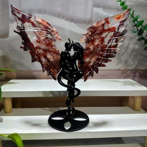 Pode incluir: Uma escultura preta de um casal abraçado, com grandes asas detalhadas em tons de vermelho, castanho e branco. A escultura é montada numa base preta com um caule giratório. As asas têm uma aparência marmoreada. A escultura é exibida numa prateleira branca.