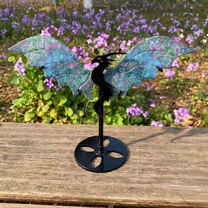May include: A black metal dragon sculpture with outstretched wings, featuring a colorful blue and pink mottled pattern. The dragon is mounted on a black stand with a circular base. The sculpture is displayed outdoors against a backdrop of purple flowers.