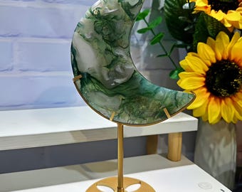 May include: A decorative crescent moon sculpture with a gold-colored stand. The moon is made of a translucent material with green and white marbling. The stand has a circular base and a slender stem. The background includes sunflowers and greenery.