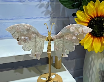 May include: A decorative butterfly sculpture with translucent wings and a gold-colored metal body and stand. The wings have a delicate, patterned appearance. The sculpture is displayed on a white surface, with sunflowers in a vase in the background.