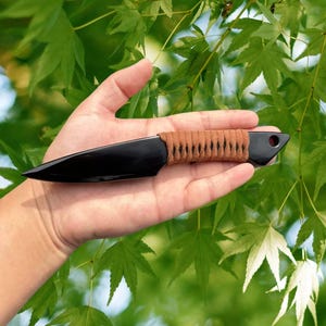 May include: A black knife with a sharp blade and a brown, wrapped handle. The knife is held in a hand against a backdrop of green leaves. The knife has a hole near the handle.